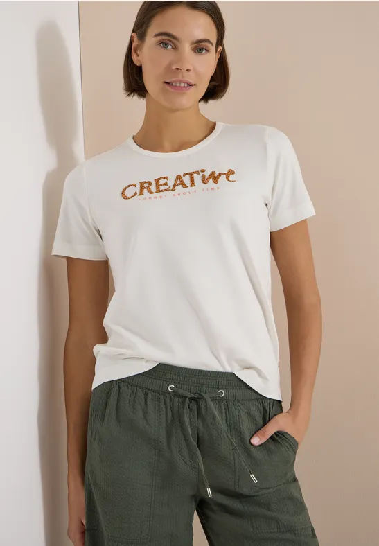 Frau in weißem T-Shirt mit 'CREATIVE'-Schriftzug und grüner Hose, eine Hand in der Tasche
