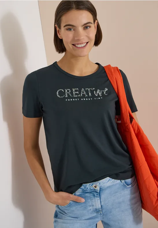Frau trägt schwarzen CREATIVE-T-Shirt, blaue Jeans und rote Tasche über der Schulter, lehnt an der Wand.