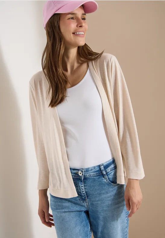 Frau in rosa Cap, weißem Top, beigem Cardigan und Jeans vor neutralem Hintergrund.