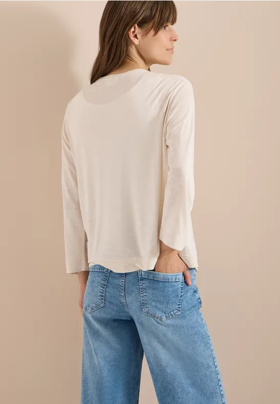 Person von hinten, trägt beiges Langarmshirt und blaue Jeans, eine Hand in der Gesäßtasche.