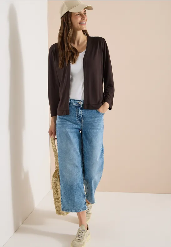 Frau in lässiger Alltagskleidung: weißes Top, schwarze Jacke, weite Jeans, Sneaker, Basecap und Strohtasche.
