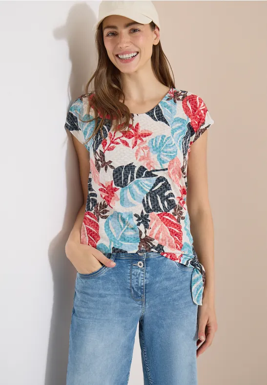 Frau trägt kurzärmeliges Top mit buntem Blumen- und Blättermuster und Knoten am Saum, kombiniert mit Jeans.