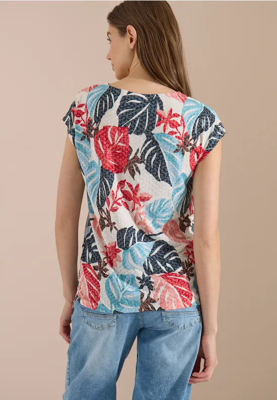 Frau von hinten trägt bunte, floral gemusterte Bluse mit kurzen Ärmeln und blaue Jeans vor neutralem Hintergrund.