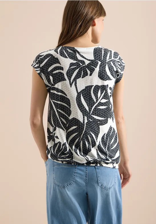 Rückansicht einer Frau in weißem Shirt mit schwarzem Blattmuster und hellblauen Jeans.