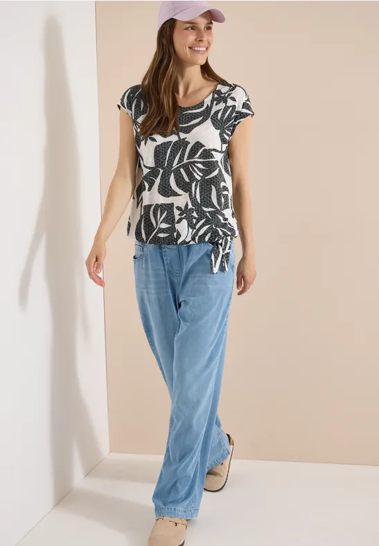 Frau trägt ein kurzärmeliges Shirt mit schwarzem Blattmuster und weite hellblaue Jeans, steht in hellem Raum.