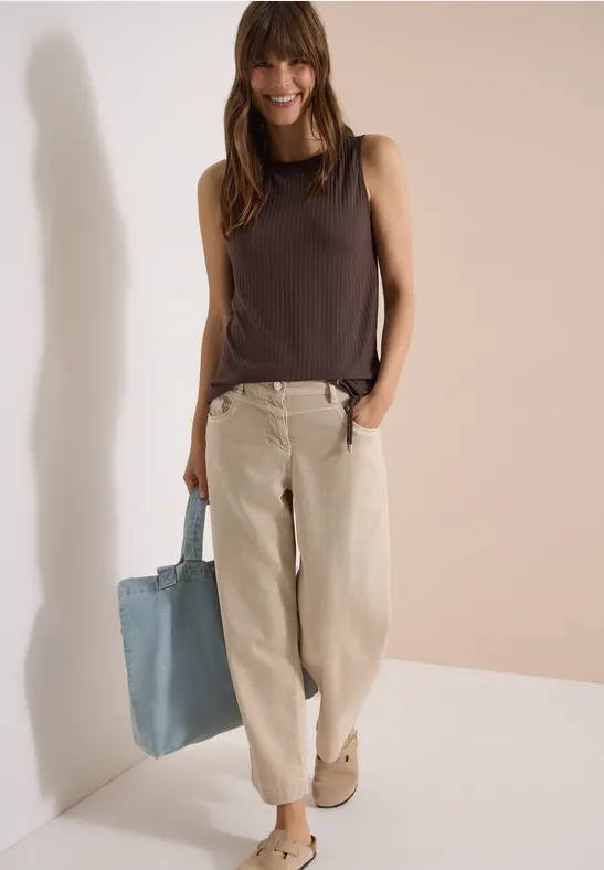 Frau trägt dunkelbraunes ärmelloses Top, helle weite Hose und blaue Stofftasche, steht vor hellem Hintergrund.