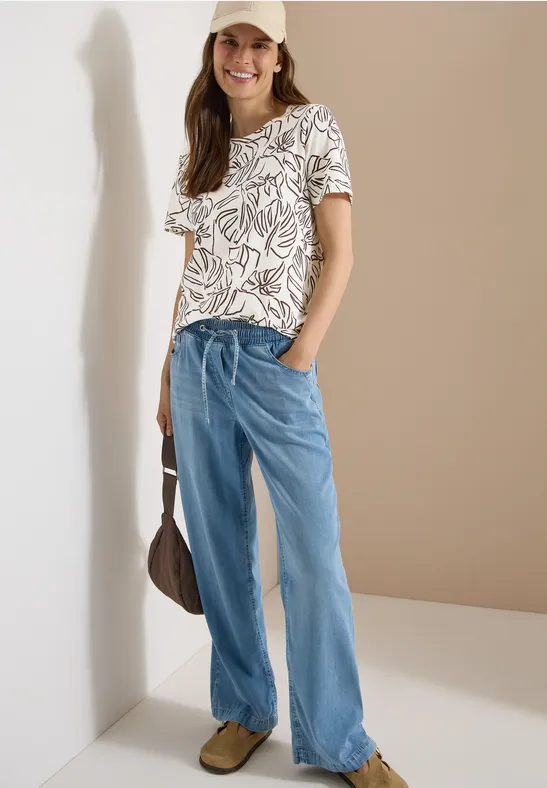 Modell trägt weißes T-Shirt mit Blatt-Print, weite hellblaue Jeans, braune Tasche und Schuhe.