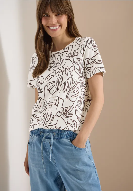 Frau in weißem T-Shirt mit Blattmuster und blauen, locker sitzenden Hose vor beigem Hintergrund.
