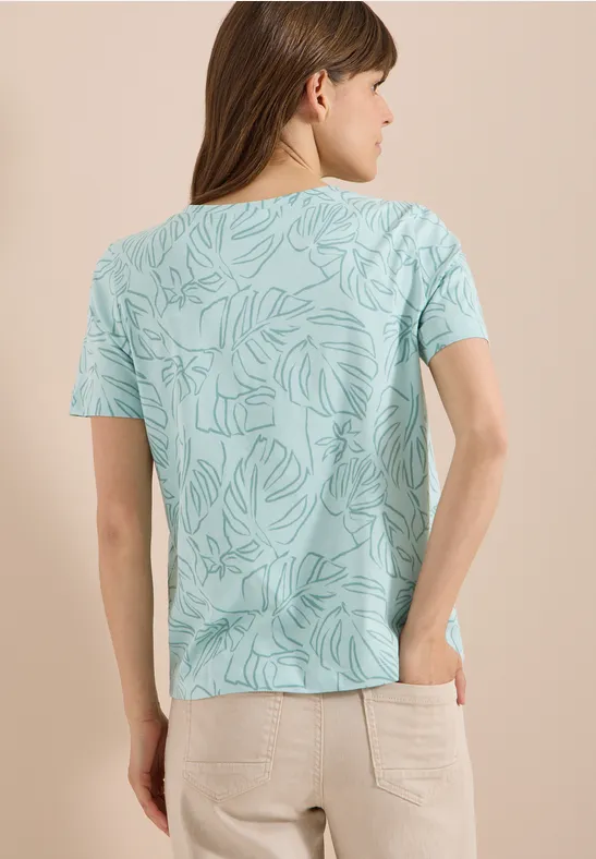 Eine Frau von hinten in mintgrünem Blattmuster-Shirt und beiger Hose.