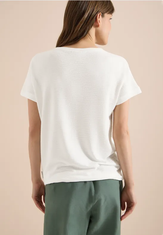 Eine Frau in weißem T-Shirt und grüner Hose, von hinten auf beigem Hintergrund.