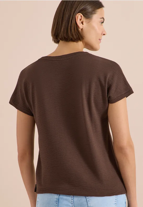 Vrouw van achteren in een bruin t-shirt en spijkerbroek voor een beige achtergrond.