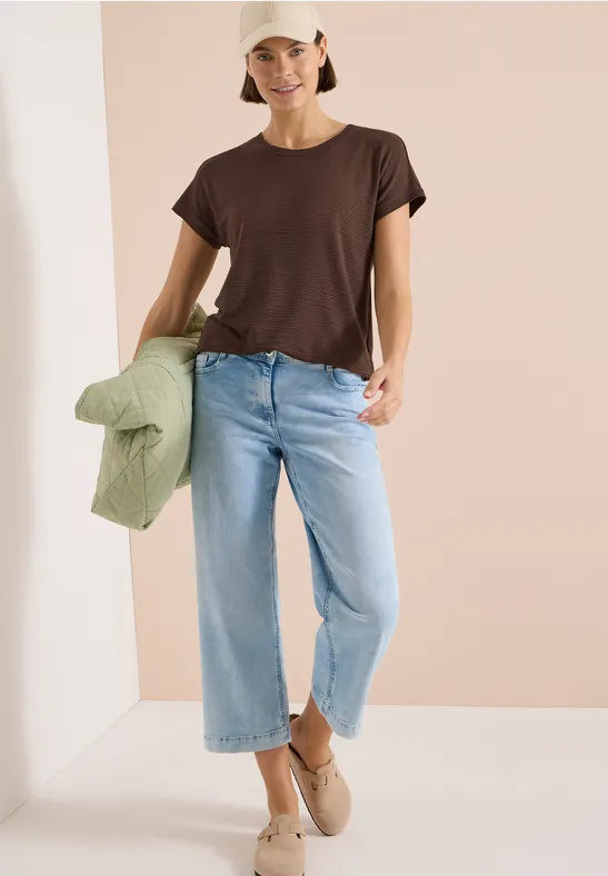Person draagt bruin T-shirt, lichte jeans en beige schoenen, houdt een groen jack in de hand.