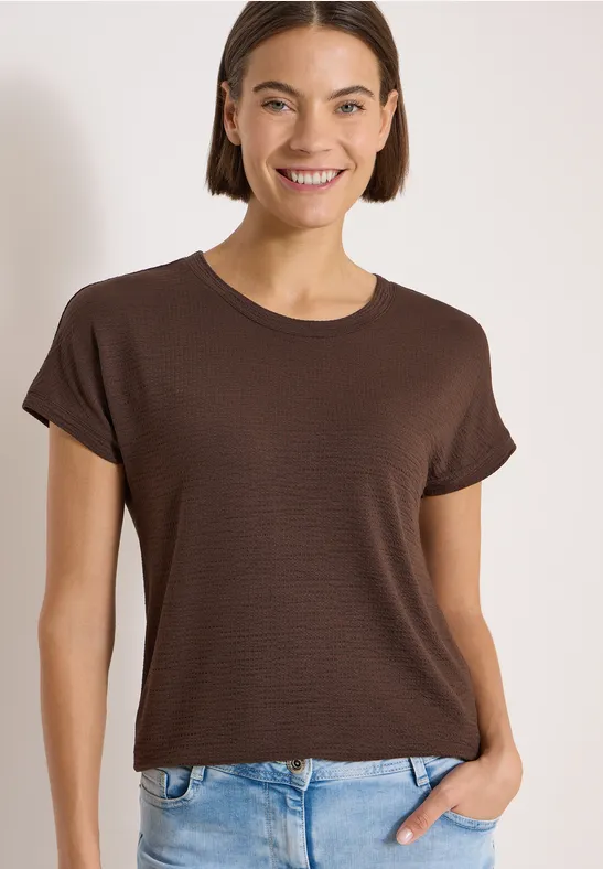 Vrouw draagt bruin t-shirt en blauwe spijkerbroek, hand in zak, tegen lichte achtergrond.