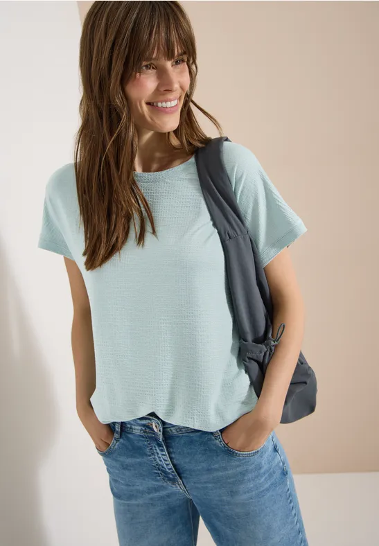 Frau trägt hellblaues T-Shirt, blaue Jeans und hängt eine dunkelgraue Tasche über die Schulter.