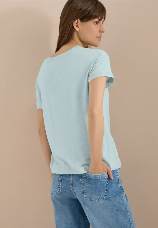 Frau von hinten in türkisfarbenem T-Shirt und blauen Jeans vor beigem Hintergrund.