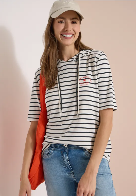 Vrouw in gestreept shirt met capuchon, spijkerbroek en rode tas voor lichte achtergrond.
