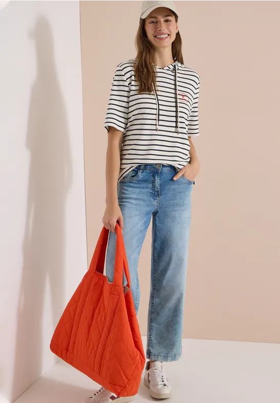 Vrouw in gestreept T-shirt, blauwe jeans en witte sneakers, met een grote oranje tas