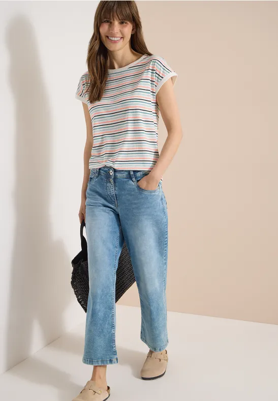 Frau lehnt an heller Wand in gestreiftem T-Shirt, blauen Jeans, Tragekorb und beigen Slippern.