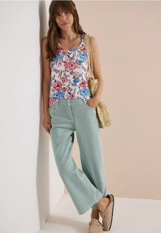 Frau lehnt lässig an Wand, trägt florales Top, helle weite Hose, Strohtasche und Sandalen.