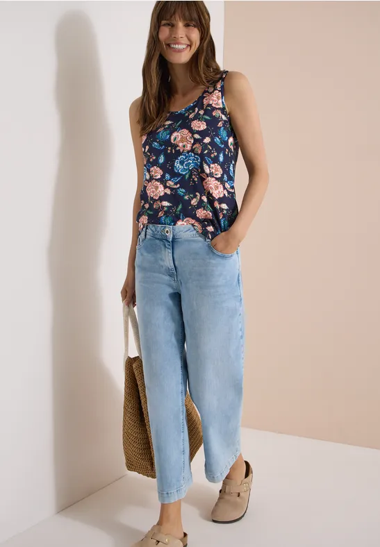 Eine Person trägt ein ärmelloses, geblümtes Top, weite Jeans, Clogs und hält eine Strohtasche.