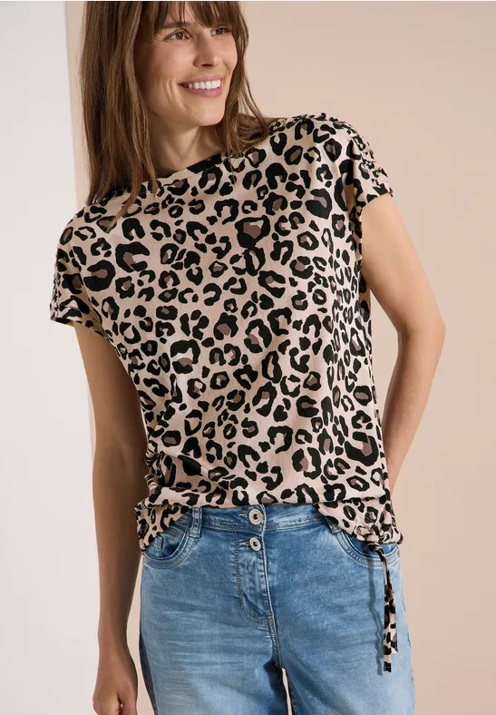 Frau in Leopardenmuster-T-Shirt mit seitlichen Bändern kombiniert mit blauer Jeans vor neutralem Hintergrund.