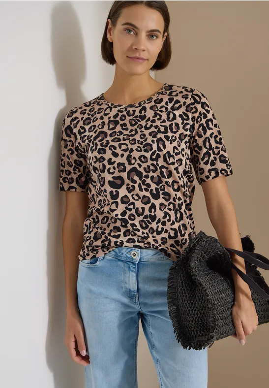 Person in beige-schwarzem Leoparden-Shirt, blauen Jeans und schwarz geflochtener Tasche vor neutralem Hintergrund.