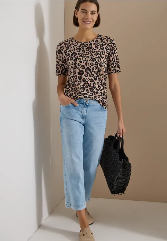 Frau in Leoparden-Shirt, hellblauen Jeans und schwarzer Flecht-Handtasche vor neutralem Hintergrund.