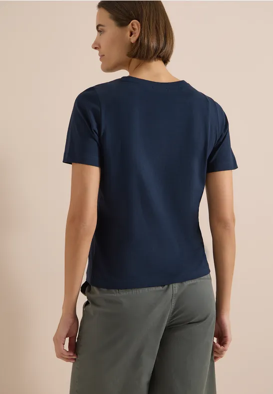 Frau von hinten in dunkelblauem T-Shirt und grauer Hose vor hellem Hintergrund.