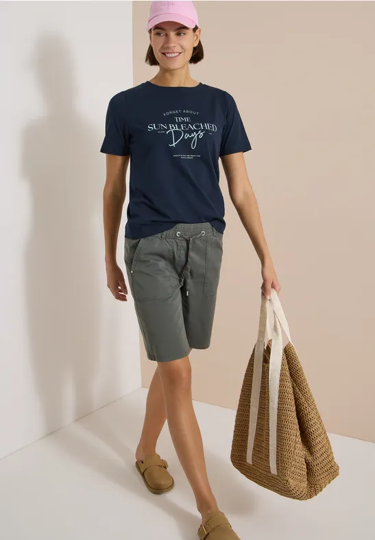 Eine Person trägt ein navy T-Shirt, graue Shorts, braune Mokassins und hält eine große geflochtene Tasche.