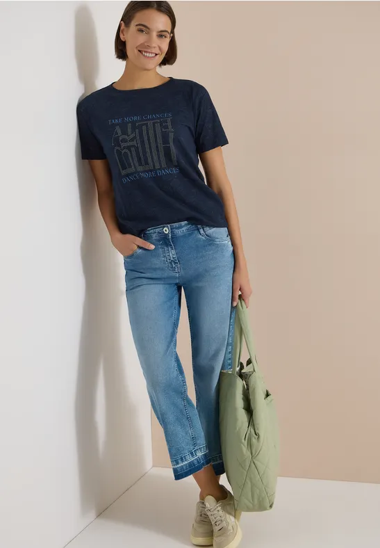 Frau lehnt lässig an der Wand in dunkelblauem T-Shirt, hellen Jeans und hält eine grüne Tasche.