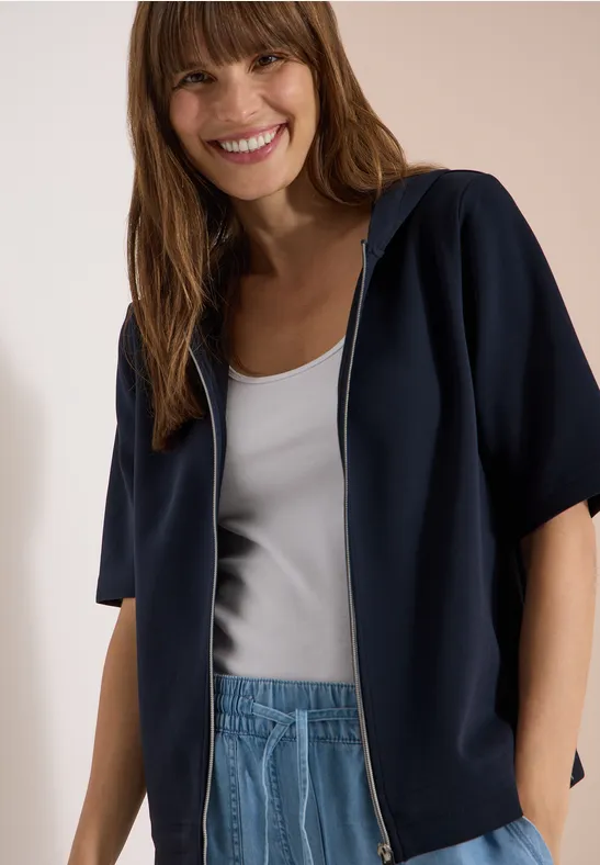 Frau in blauem Kurzarm-Hoodie, weißem Top und Jeanshose, Gesicht unscharf.