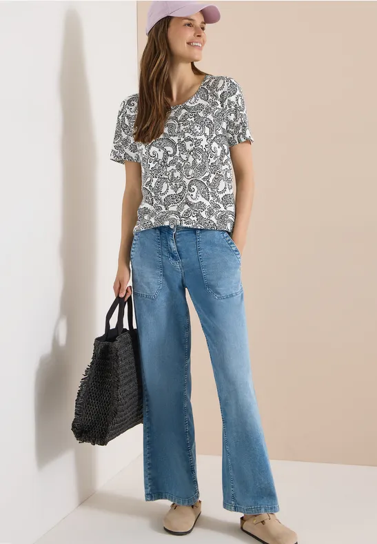 Frau in gemustertem T-Shirt, weitem Jeans, Basecap und geflochtener Tasche