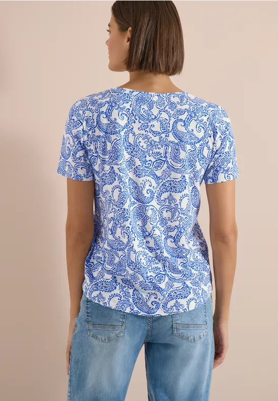 Rückansicht einer Frau in kurzem, blau-weiß gemustertem T-Shirt und hellblauen Jeans vor beigem Hintergrund.