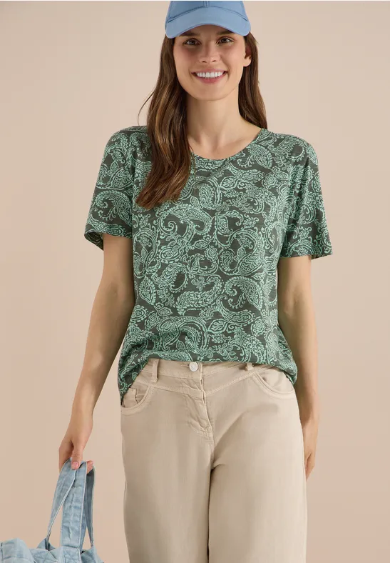Frau trägt grünes, gemustertes T-Shirt, helle Hose und hält eine hellblaue Tasche.