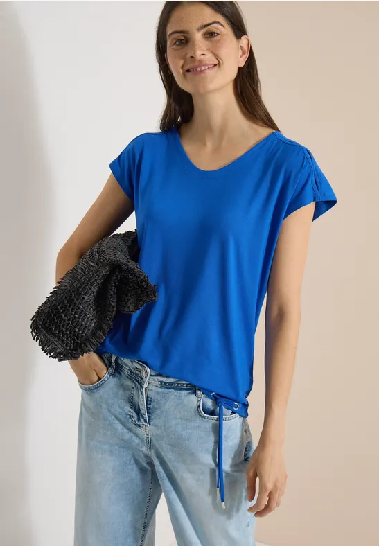 Femme portant un T-shirt bleu à cordon, jean clair et sac noir texturé à l’épaule, fond uni clair.