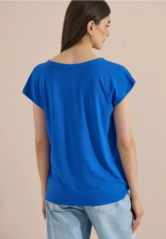 Femme de dos portant un T-shirt bleu ample et un jean clair sur fond neutre.