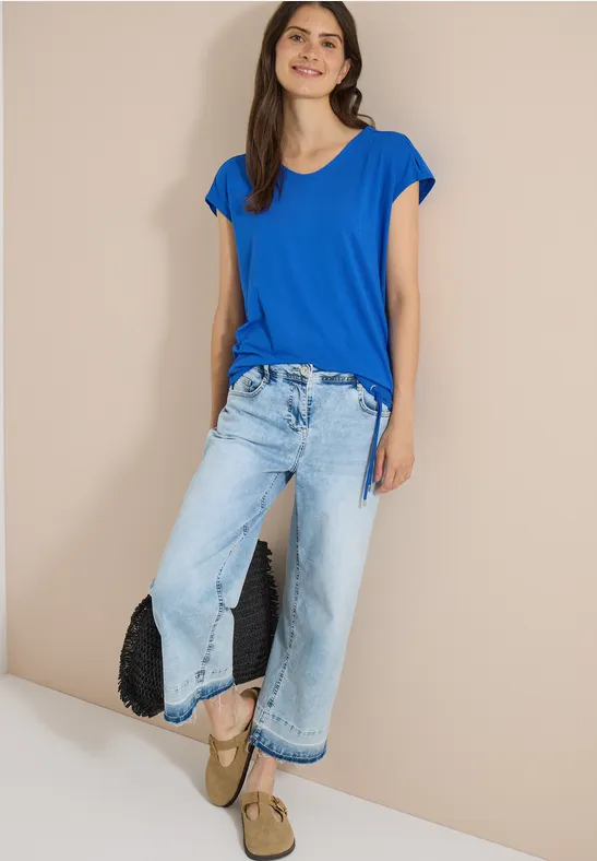 Femme en T-shirt bleu, jean large, sandales à plateforme et pochette noire devant un mur beige.