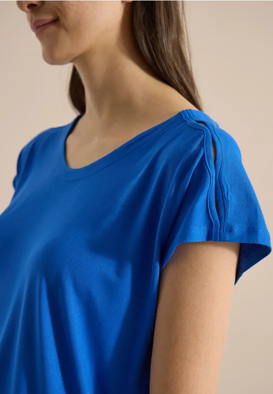 Une femme de profil porte un t-shirt bleu vif à col rond avec des manches courtes légèrement froncées.