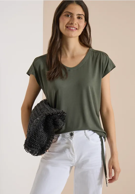 Een vrouw draagt een groen T-shirt en witte jeans, ze houdt een zwarte netzak voor een neutrale achtergrond.