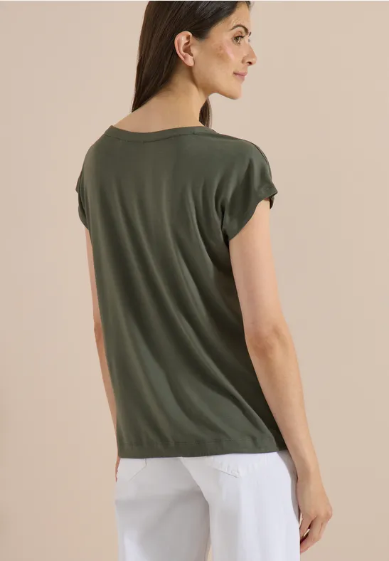 Rugaanzicht van een vrouw in een groen T-shirt met omgeslagen mouwen en witte broek tegen een lichte achtergrond.
