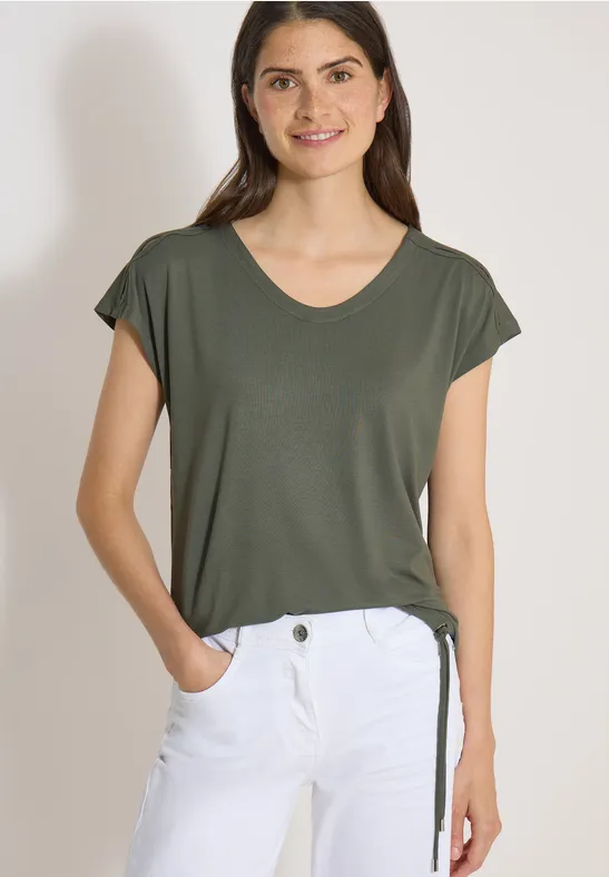 Vrouw in olijfgroene T-shirt met korte mouwen en witte broek.