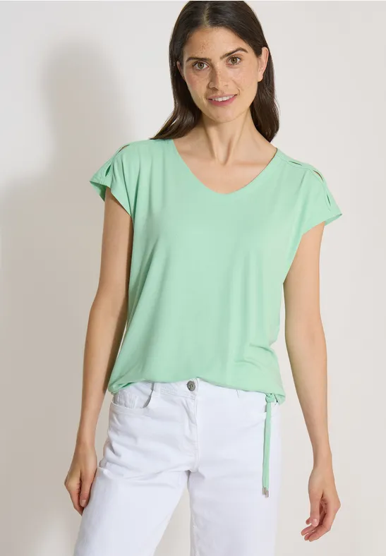 Modèle porte un T-shirt vert clair à manches courtes et un pantalon blanc sur fond neutre.