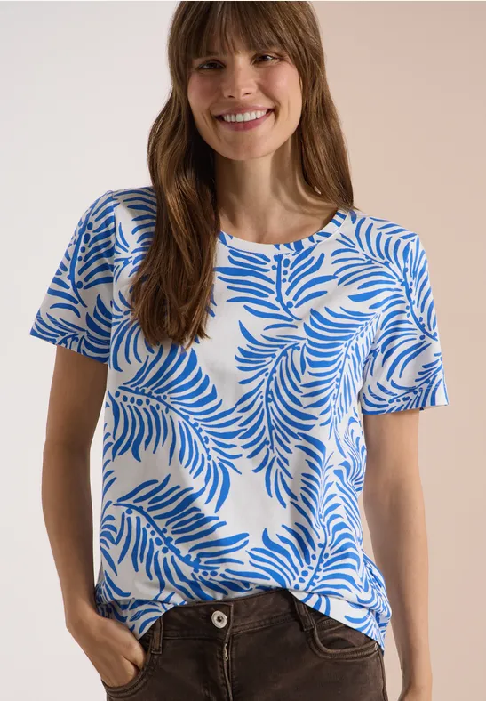 Frau trägt weiß-blaues T-Shirt mit Palmenblättermuster, steht mit Händen in den Taschen