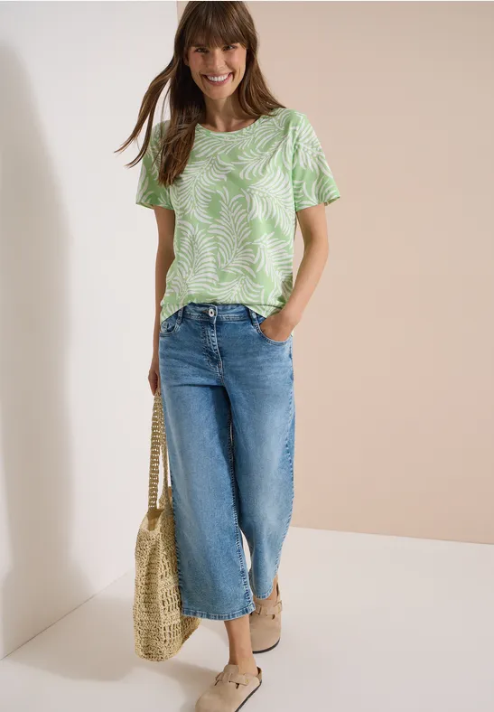 Frau in grün-weiß gemustertem T-Shirt, weiten Jeans-Culottes, Basttasche und Sandalen.