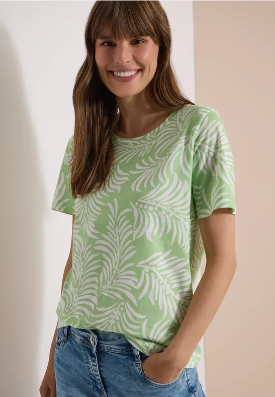 Frau mit grünem T-Shirt mit weißem Blättermuster und Jeans, Hände in den Taschen.