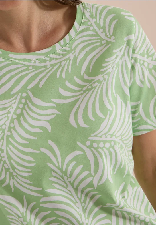 Frau in grünem T-Shirt mit weißem, organischem Palmenmuster, nur Oberkörper zu sehen.