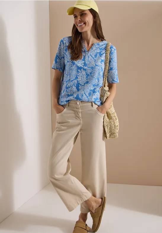 Vrouw in blauw bedrukt T-shirt met korte mouwen, ruime beige broek, rieten tas en loafers voor een neutrale achtergrond.