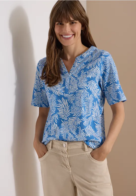 Vrouw in blauw-wit gebloemd shirt met korte mouw en lichte broek, handen in de zakken, voor beige achtergrond.