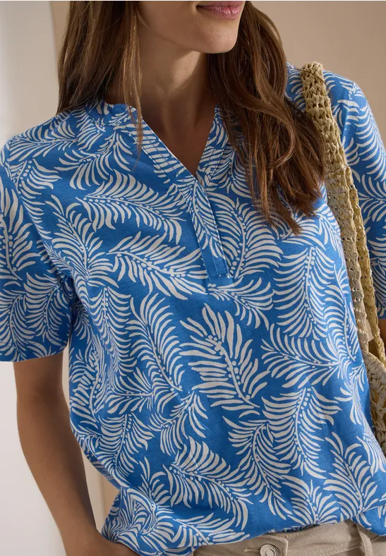 Vrouw in blauw-wit palmbladprint blouse met V-hals en korte mouwen, schoudertasriem zichtbaar.