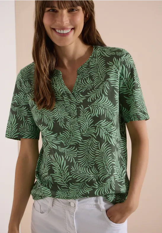 Femme portant un t-shirt vert à motif feuillage et un pantalon blanc, mains dans les poches.